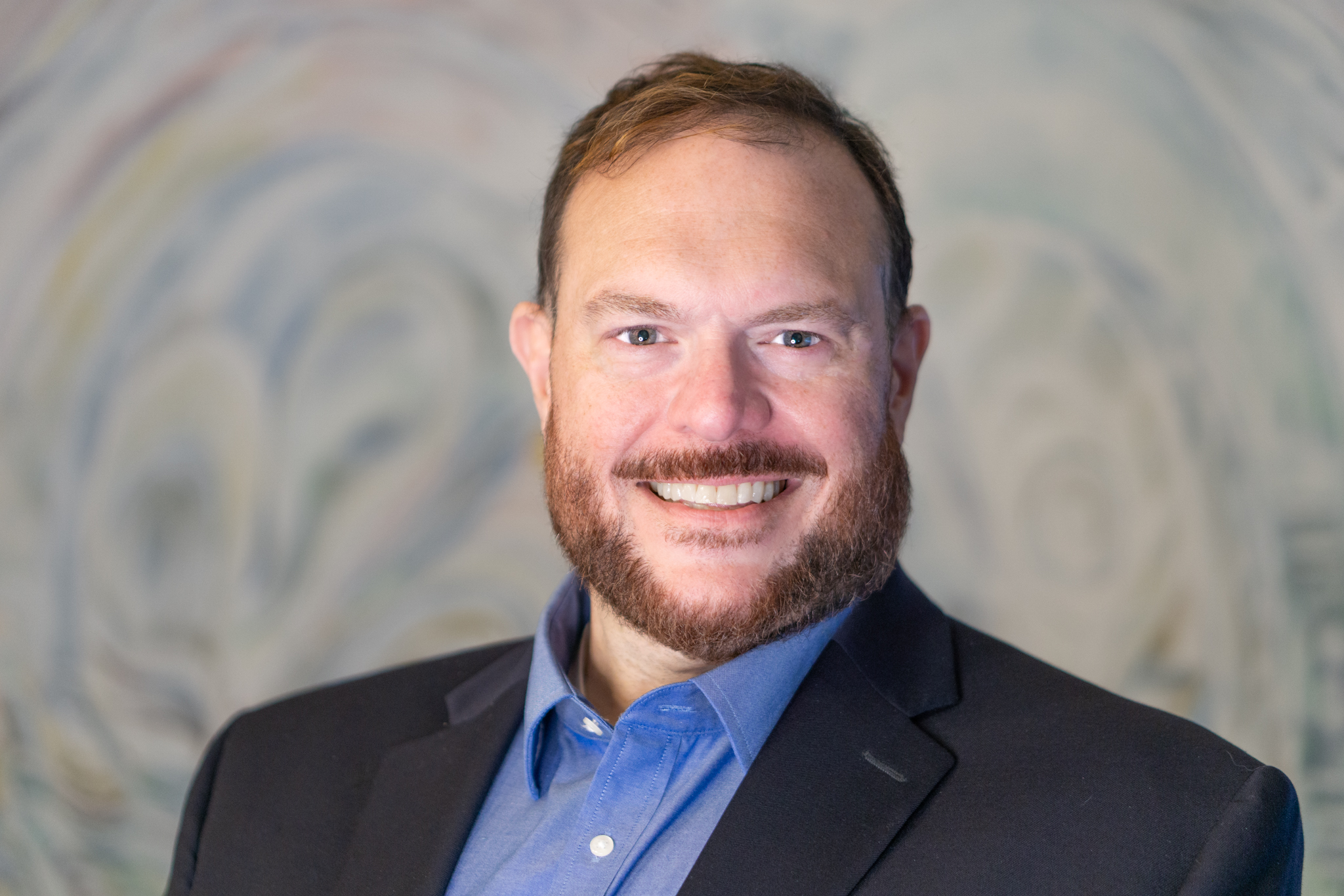 Robert K. Arena, Jr. - professional headshot. A man with short brown hair and a warm smile, wearing a blue dress shirt, photographed outdoors.